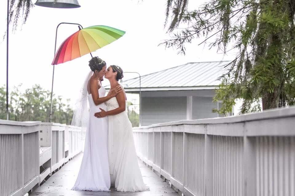 Two brides kissing on bridge under rainbow umbrella