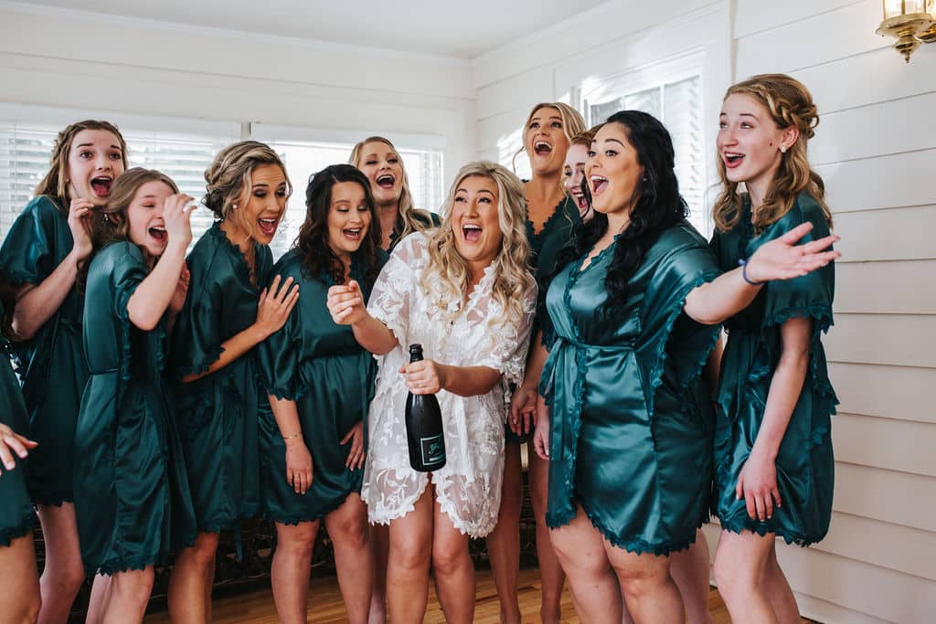 Bride opening bottle of champagne surrounded by bridesmaids.